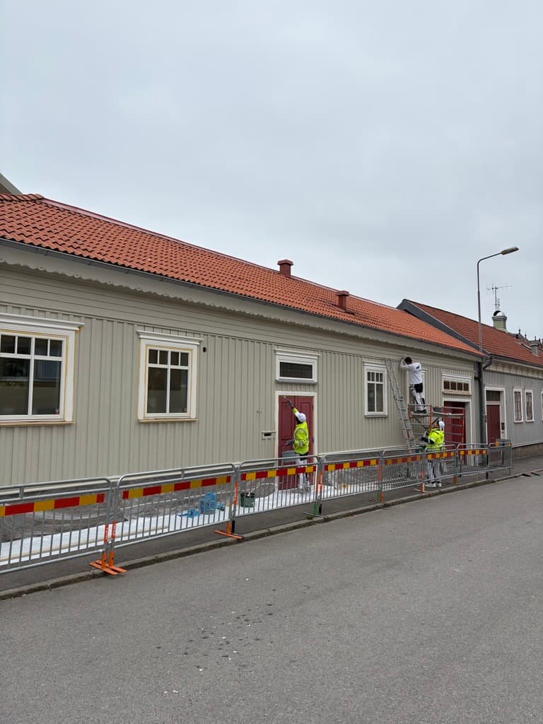 Ljus träfasad med rött tegeltak under ommålning, avspärrad mot gatan.